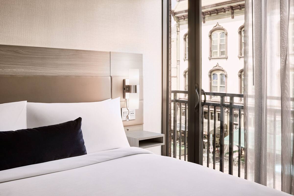 Ein Hotelzimmer mit einem Bett, einem Nachttisch, einer Lampe und Blick auf ein Gebäude durch ein Fenster.