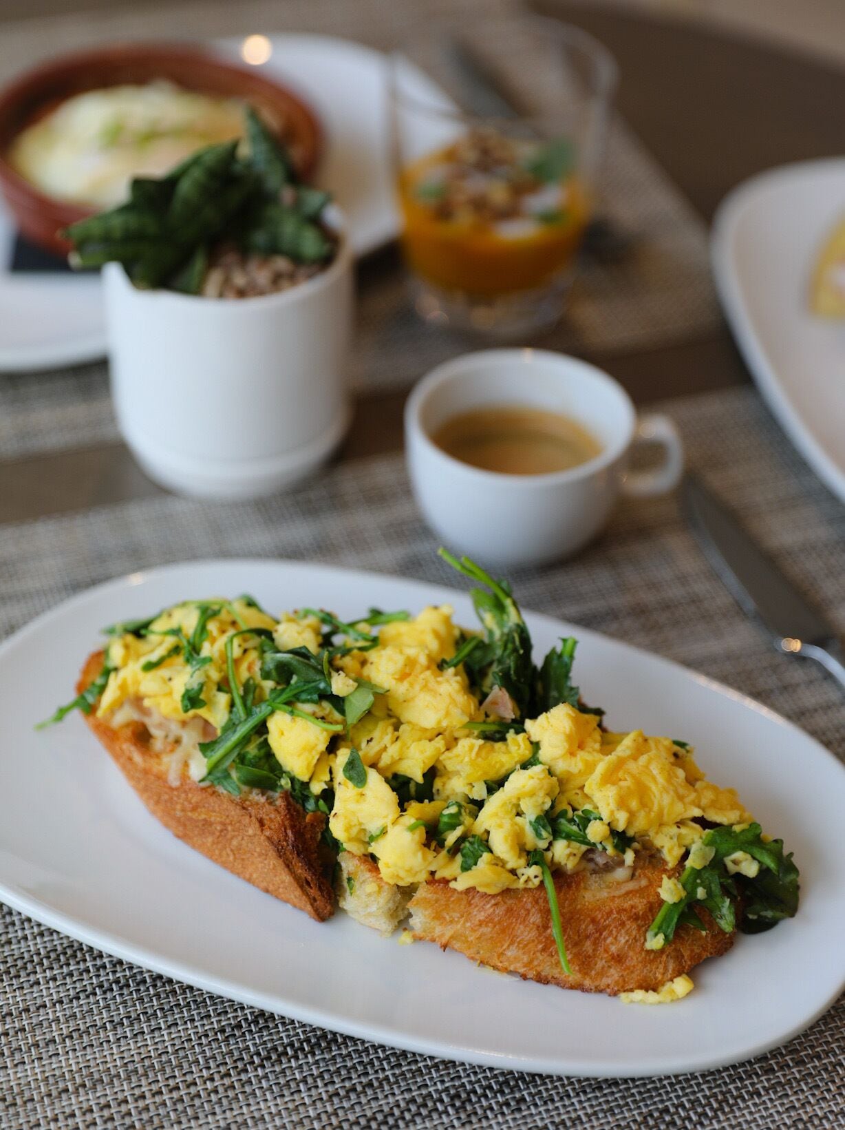 Un plato de huevos revueltos sobre tostadas, hojas verdes, una taza de café y un tazón de sopa sobre una mesa.
