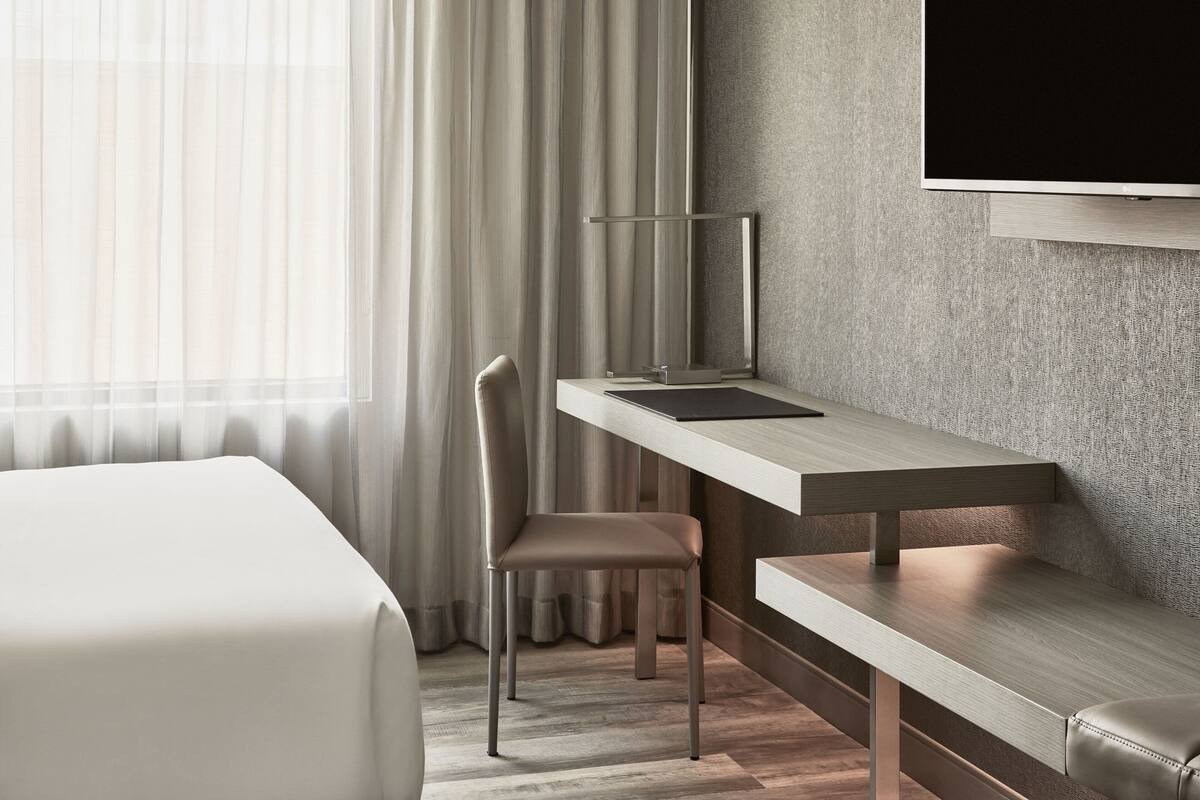 Ein modernes Hotelzimmer mit einem Bett, einem Schreibtisch mit Stuhl, einem Flachbildfernseher und einem großen Fenster mit Vorhängen.