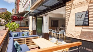 An outdoor seating area with wooden benches and cushions, a covered patio, and a sign for AC Hotels Marriott. - AC Hotel by Marriott San Diego Downtown Gaslamp Quarter (San Diego)