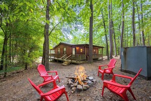Une cabane entourée d’arbres, avec un foyer et des chaises rouges devant.