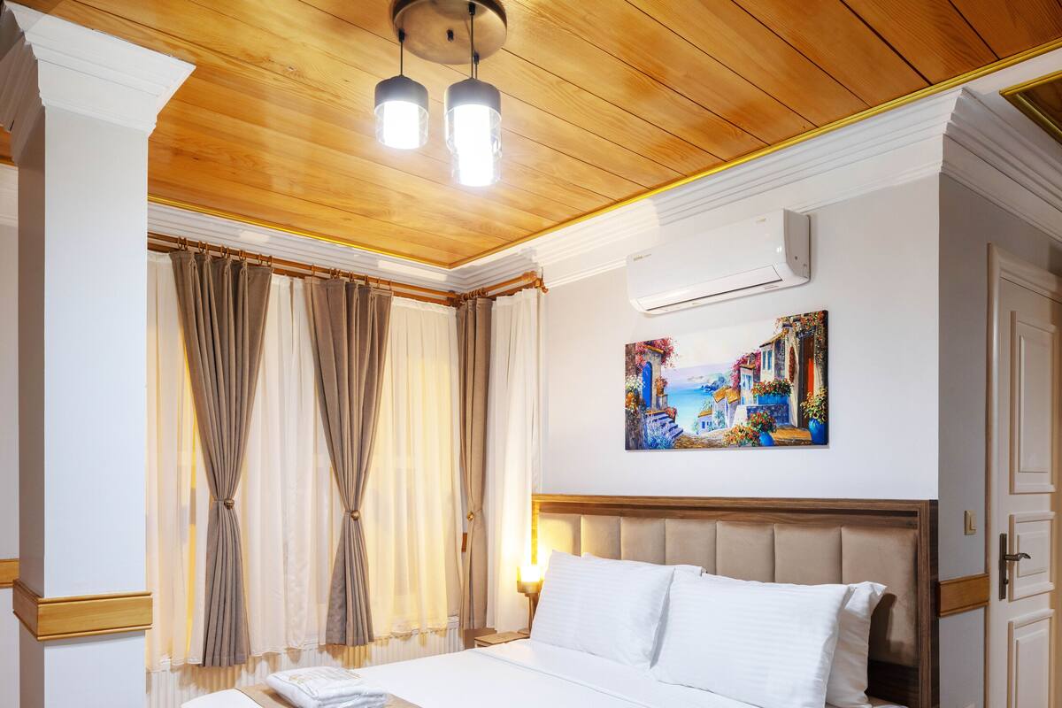 A hotel room with a large bed, wooden flooring, a painting on the wall, and a ceiling with wooden beams and pendant lights.