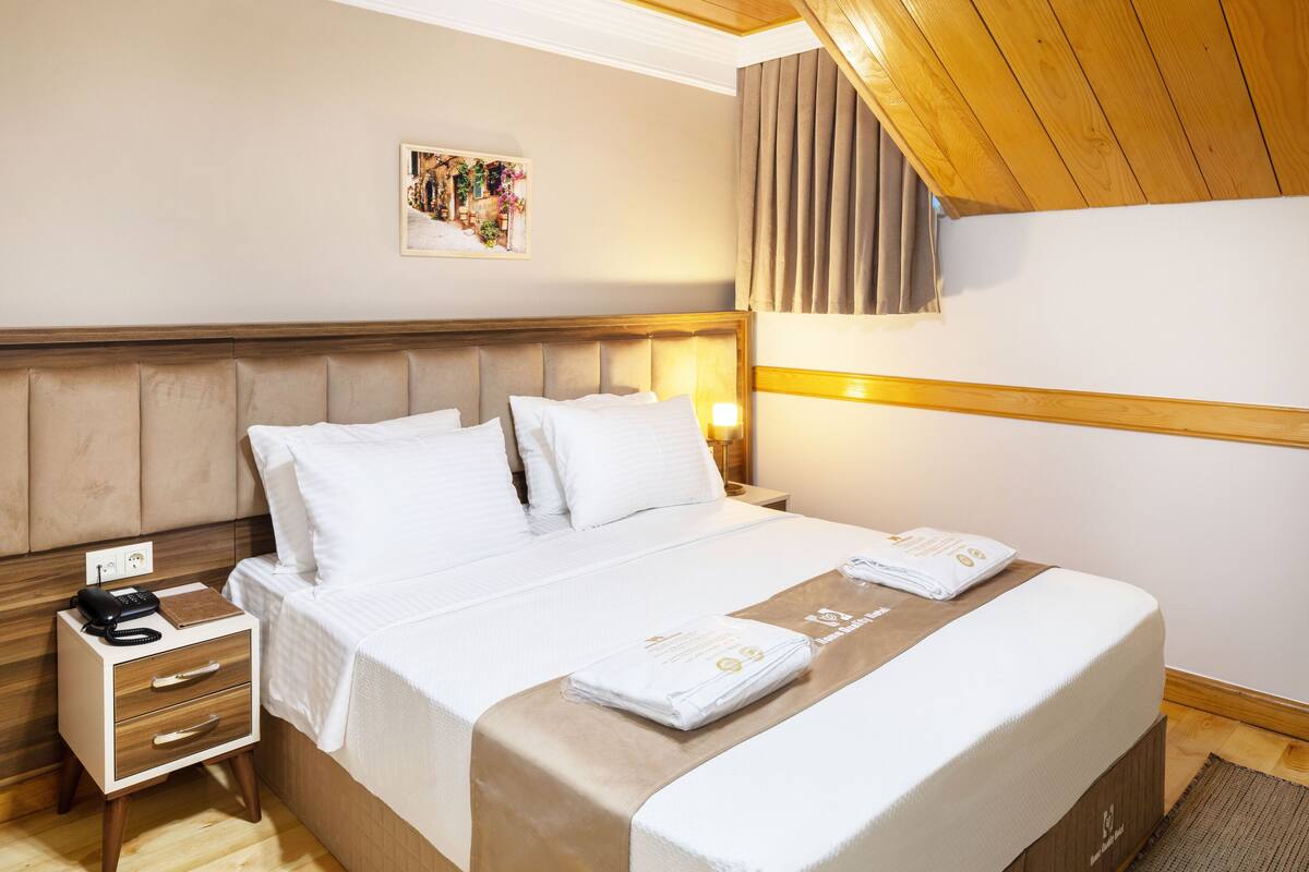 A hotel room with a wooden ceiling, a bed with white linens, a nightstand with a phone, and a wall-mounted lamp.