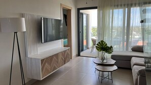 Ein modernes Wohnzimmer mit einem an der Wand befestigten Flachbildfernseher, einem Deckenventilator, einer Schiebeglastür, die auf einen Balkon führt, und einer Topfpflanze auf einem kleinen Tisch.