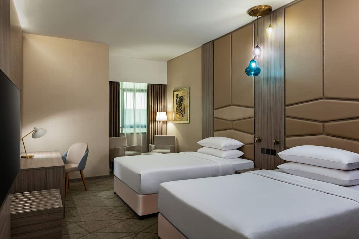 A hotel room with two beds, a desk, a chair, a TV, and a large window with curtains.
