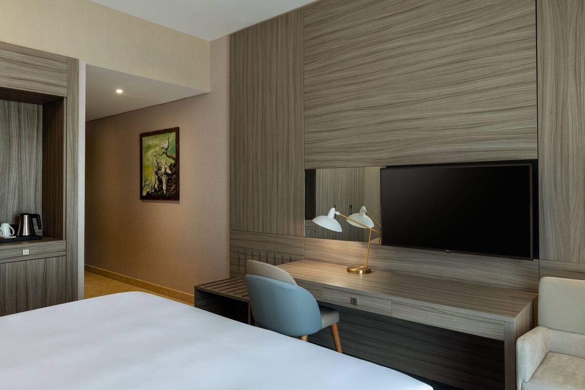 A hotel room with a large bed, a desk with a chair, a flat-screen TV, a coffee machine, and a painting on the wall.