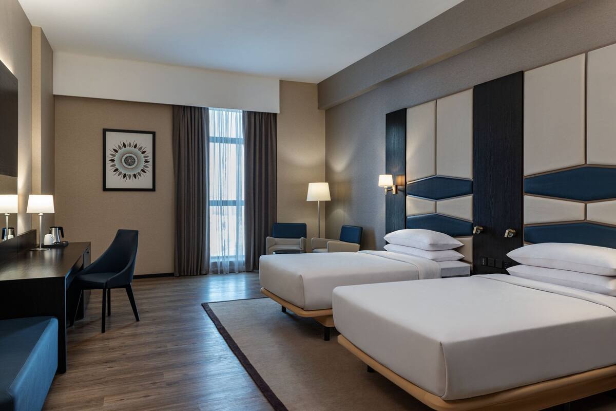 A hotel room with two beds, a desk, a chair, and a large window with curtains.