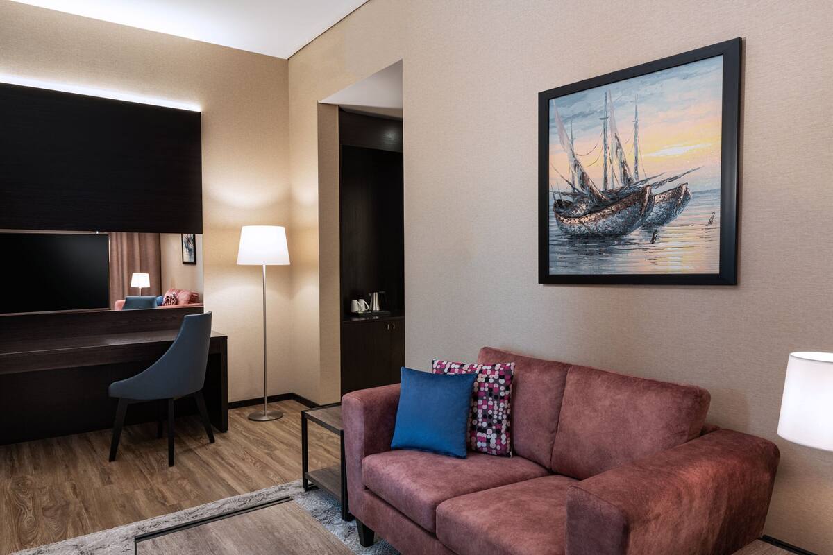 A modern hotel room with a dark wood desk, a blue chair, a pink sofa, and a framed painting of a boat on the wall.