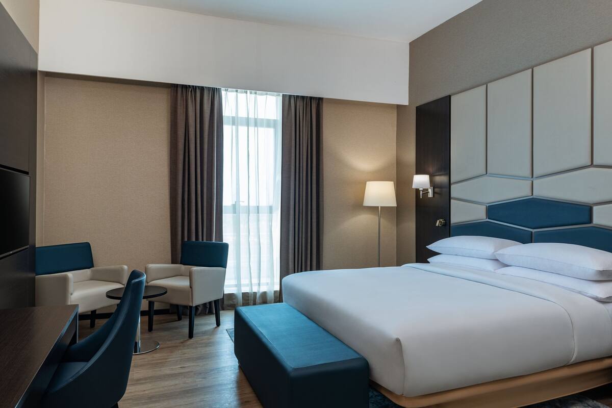 A hotel room with a bed, two armchairs, a small table, a desk, and a TV.