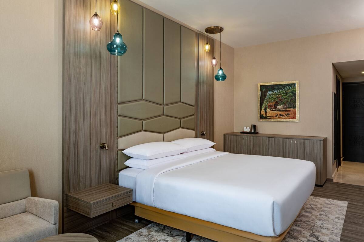 A modern bedroom with a large bed, a wooden headboard, a nightstand, and a bedside table. There is a painting on the wall and a door to the right.