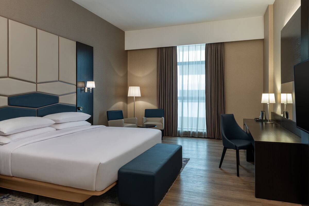 A hotel room with a large bed, two bedside tables, a desk with a chair, a TV, and a window with curtains.