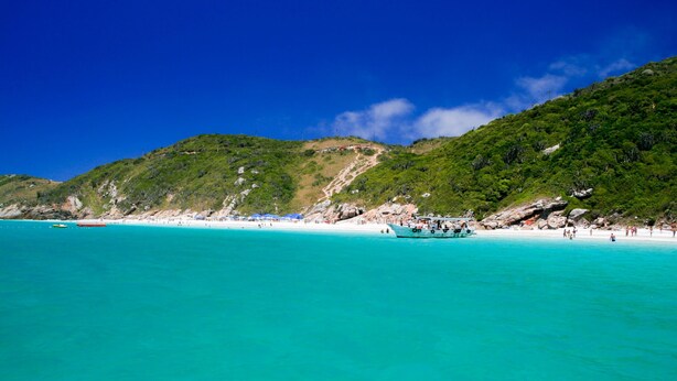 A beach with clear turquoise water, a boat, and a hilly landscape in the background. - Recanto das Tartarugas Aluguel Economico (Arraial do Cabo)