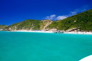 Een strand met helder turquoise water, een boot en een heuvelachtig landschap op de achtergrond.
