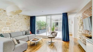 Un salon moderne avec un mur en pierre, un canapé gris, une table basse sur laquelle est posé un jeu d’échecs, et une porte-fenêtre coulissante donnant sur un espace extérieur.