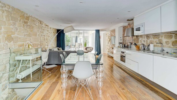 Une cuisine moderne avec des meubles blancs, un mur en pierre et un coin repas comprenant une table et des chaises en verre.