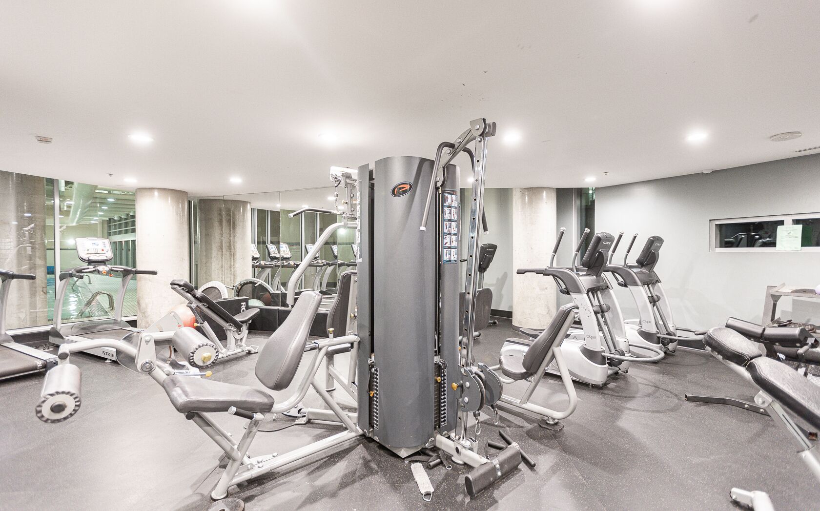 A gym with various exercise machines, including treadmills and weight stations, arranged in a spacious room with large windows.