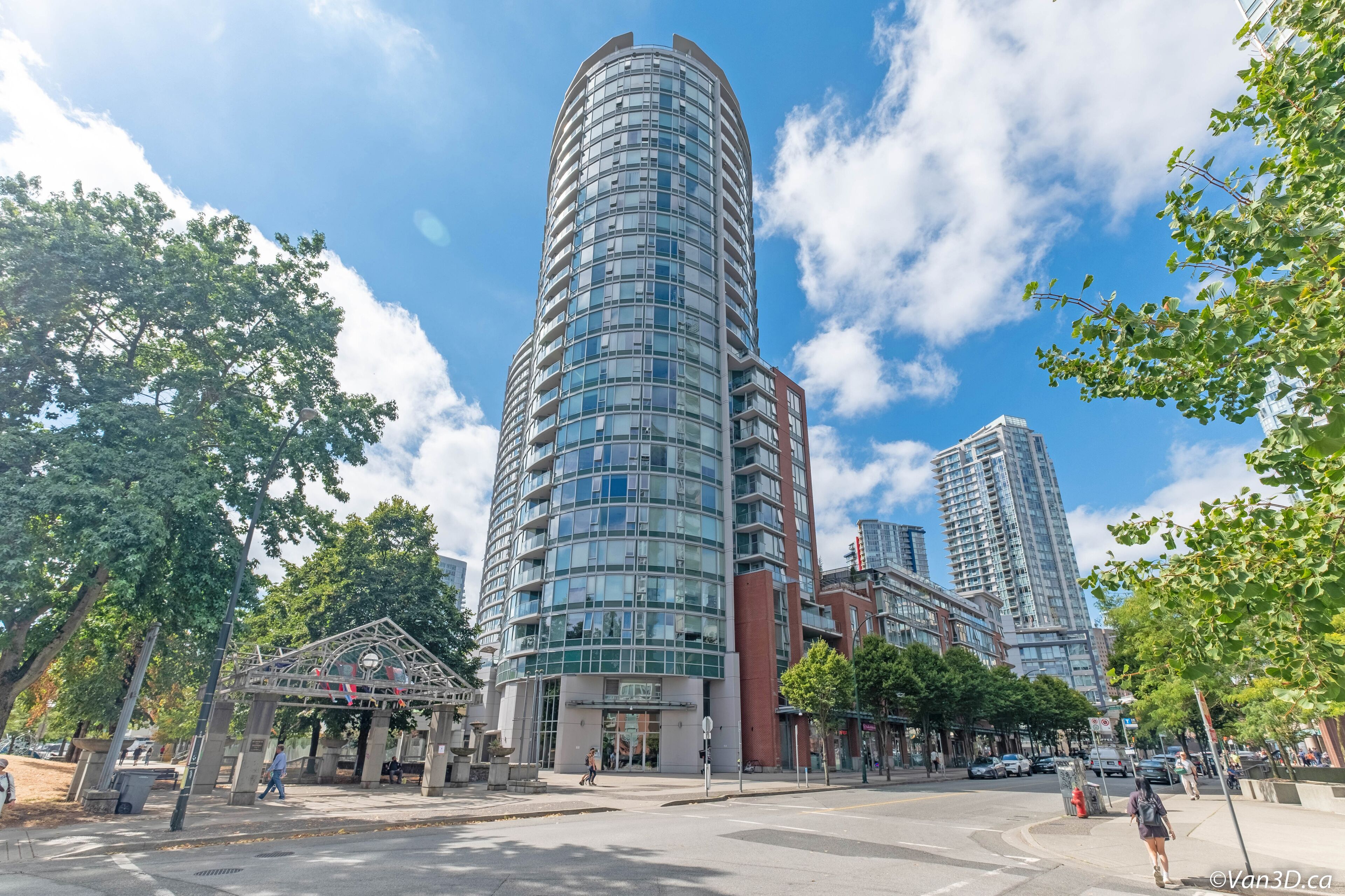 A high-rise building with a curved glass facade, a pedestrian entrance, and a street view with trees and other buildings.