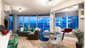 Un salon moderne doté d’une grande fenêtre offrant une vue sur la ville, d’un coin salon confortable avec un canapé et des fauteuils, et d’une table à manger avec des chaises.