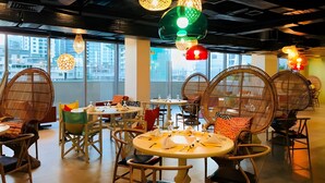 Un espace de restauration moderne avec des tables rondes et des chaises en osier, de grandes fenêtres offrant une vue sur la ville et des luminaires suspendus au design original.