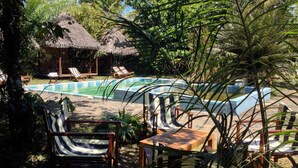 Área de piscina com espreguiçadeiras e uma estrutura com telhado de palha.