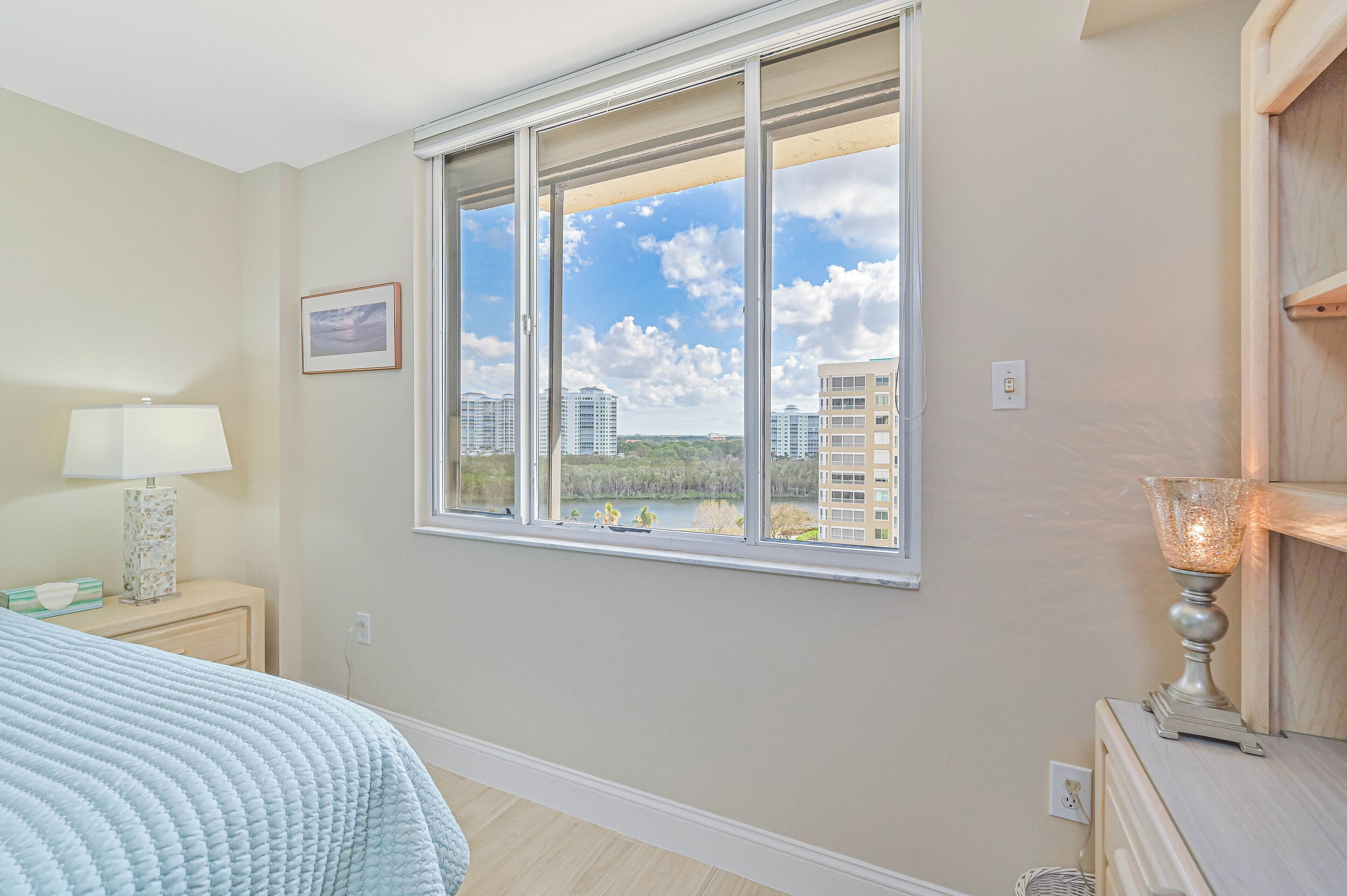 A bedroom with a large window, a bed, a nightstand, a lamp, and a dresser.