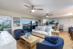 A spacious living room with a large sofa, two armchairs, a coffee table, and a ceiling fan. There are paintings on the walls and a view of the ocean through the windows. - 3BR Ka’anapali Retreat | Ocean Views, Gorgeous renovation, Walk to Beach (Lahaina)