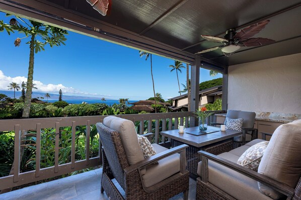 A covered outdoor seating area with wicker furniture, a table, and a view of the ocean and palm trees. - 3BR Ka’anapali Retreat | Ocean Views, Gorgeous renovation, Walk to Beach (Lahaina)
