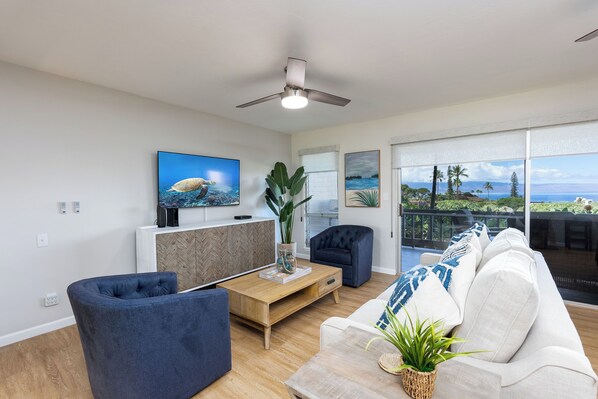 A modern living room with a sofa, a coffee table, and a TV. There is a balcony with a view of the ocean.