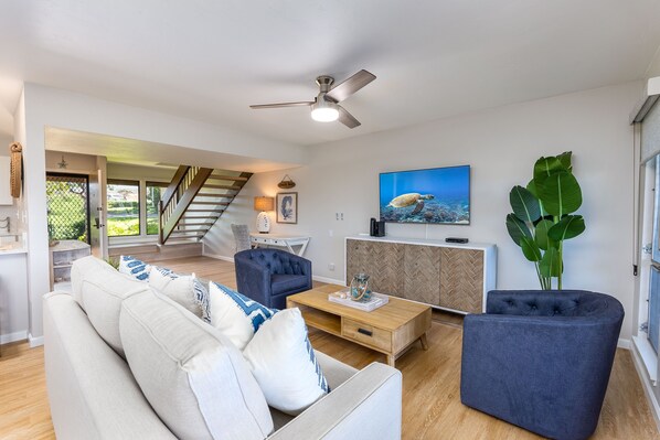 A modern living room with a sofa, armchairs, a coffee table, and a TV. There is a staircase leading to another level.