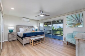 A bedroom with a bed, bedside tables, a bench, a ceiling fan, a large window, and a balcony with a view of greenery.