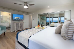 A bedroom with a bed, a ceiling fan, a television displaying a turtle, and a balcony with a view of the ocean. - 3BR Ka’anapali Retreat | Ocean Views, Gorgeous renovation, Walk to Beach (Lahaina)