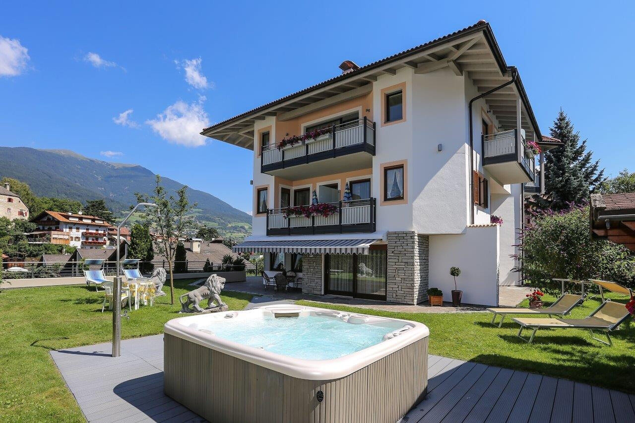 A two-story building with a balcony, a hot tub, and a patio area with chairs and a statue.