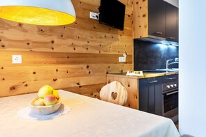 Un intérieur en bois comprenant une table à manger, un saladier rempli de fruits, une chaise en forme de cœur, un coin cuisine avec une cuisinière et un téléviseur fixé au mur.