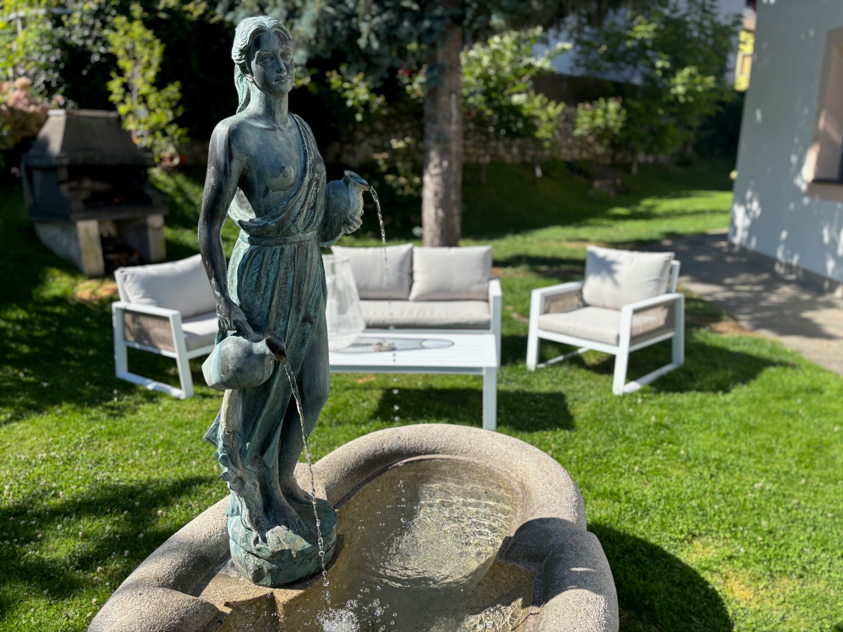 Eine Statue einer Frau, die eine Kanne hält, ein Steinfountain und weiße Gartenmöbel in einer Gartenanlage.