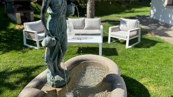 Eine Statue einer Frau, die eine Kanne hält, ein Steinfountain und weiße Gartenmöbel in einer Gartenanlage.