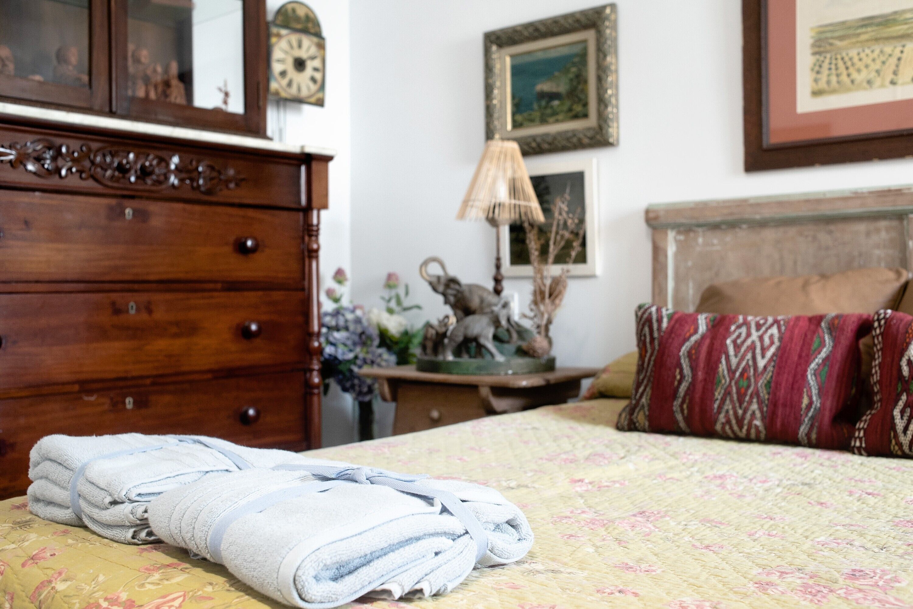 A neatly made bed with a floral bedspread, a wooden dresser with drawers, a lamp, and a framed picture on the wall.