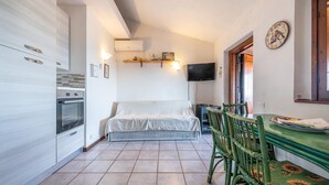 A compact kitchen with a built-in oven, a dining area with a table and chairs, a sofa, and a TV mounted on the wall. - Holiday Home 'Bianca' with Sea View, Private Terrace and Air Conditioning (Costa Rei)
