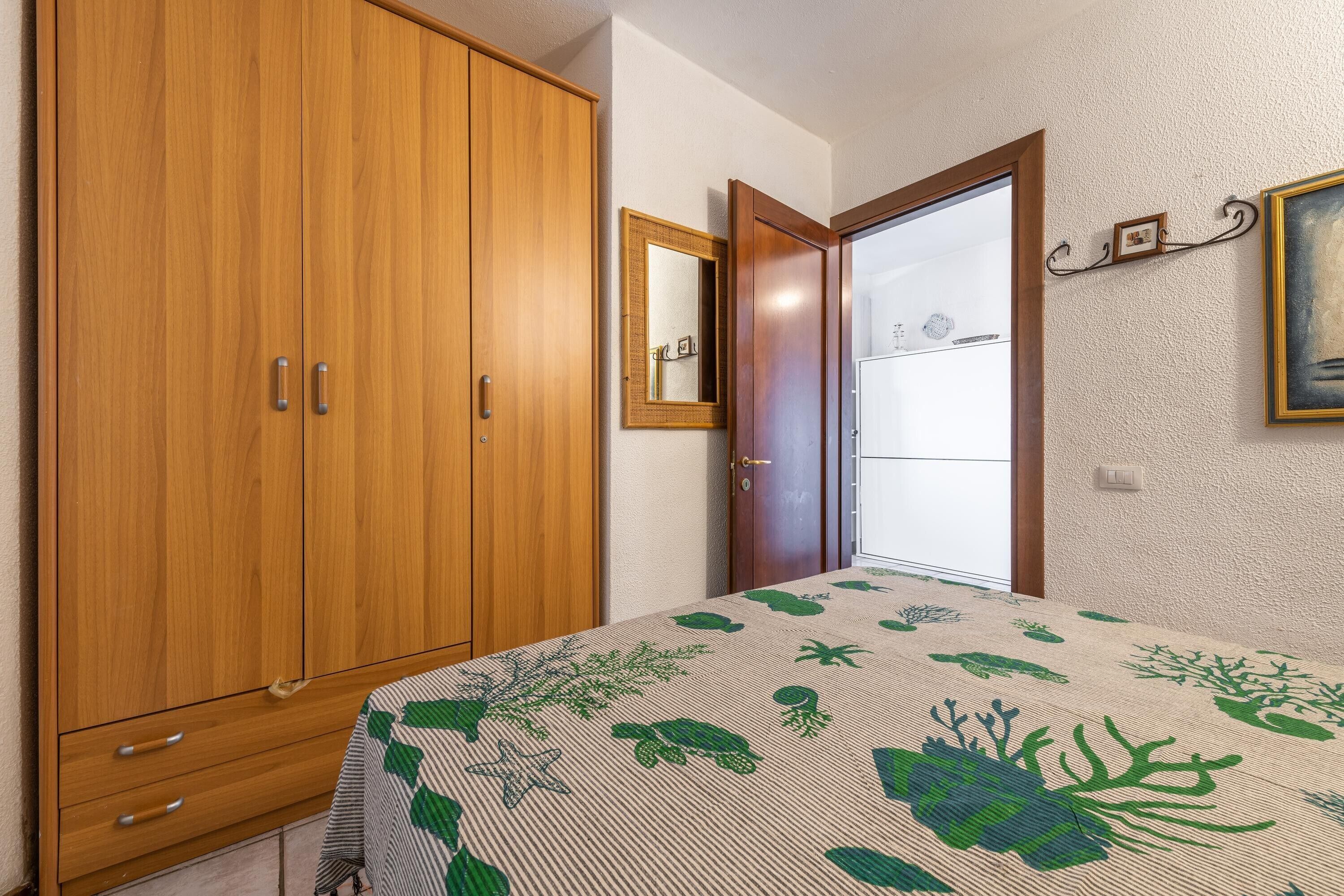 Un dormitorio con un armario de madera, una cama con colcha estampada, un espejo y una puerta que da a otra habitación.