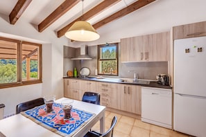 A modern kitchen with wooden beams, a dining table with a patterned tablecloth, and a view of greenery outside.