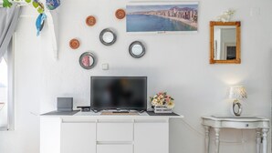 Et moderne værelse med et hvidt skrivebord, et fladskærms-TV, dekorative spejle, et billede af en by og et lille bord med en lampe.