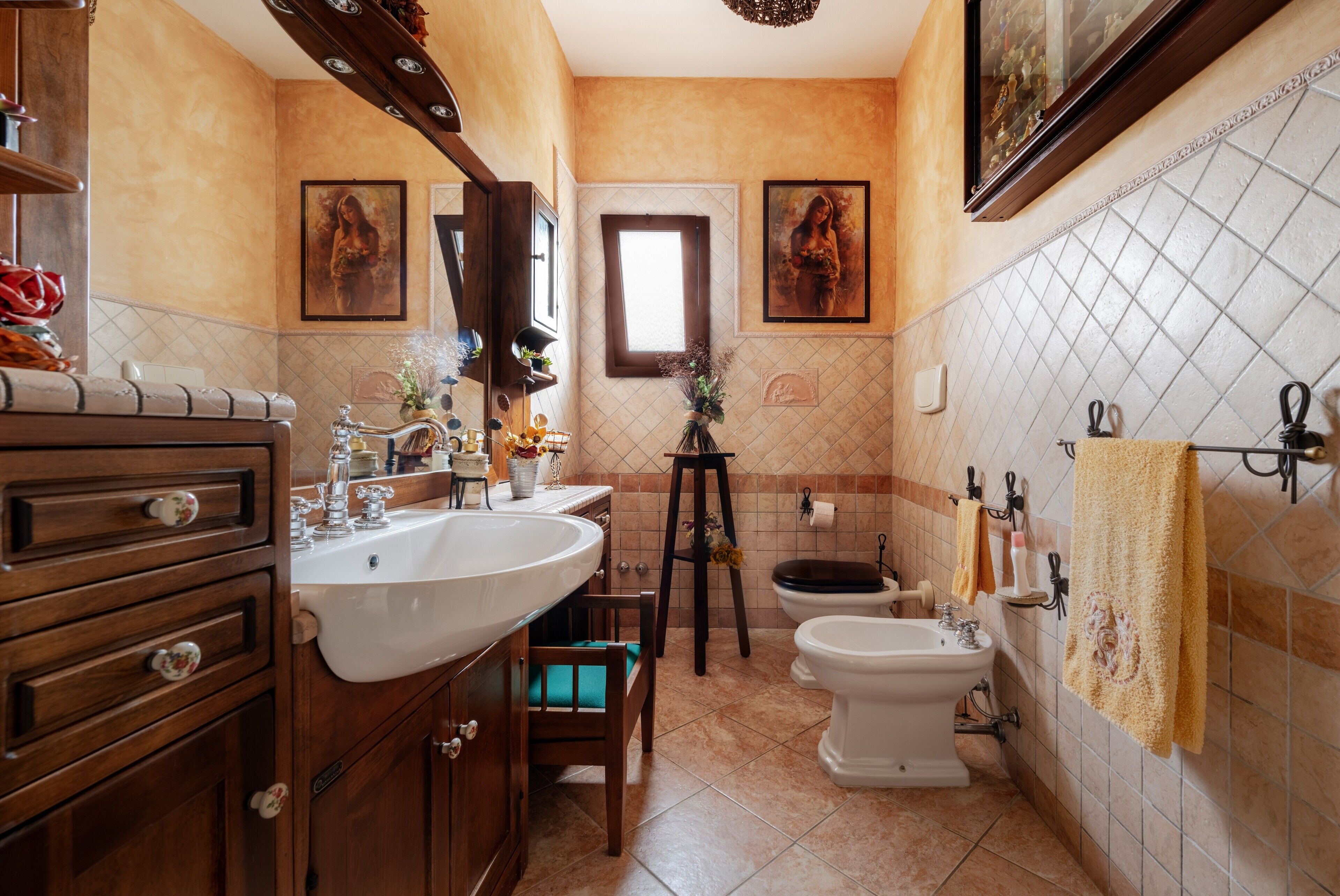 A bathroom with a wooden vanity, a sink, a toilet, and a bidet. There are framed pictures on the wall and a window above the toilet.