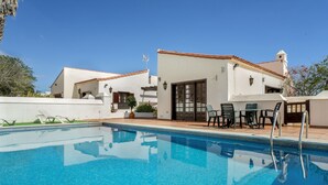 En swimmingpool med klar, blå vandoverflade, omgivet af en hvid bygning med et flis tag, og en terrasse med udendørs møbler.