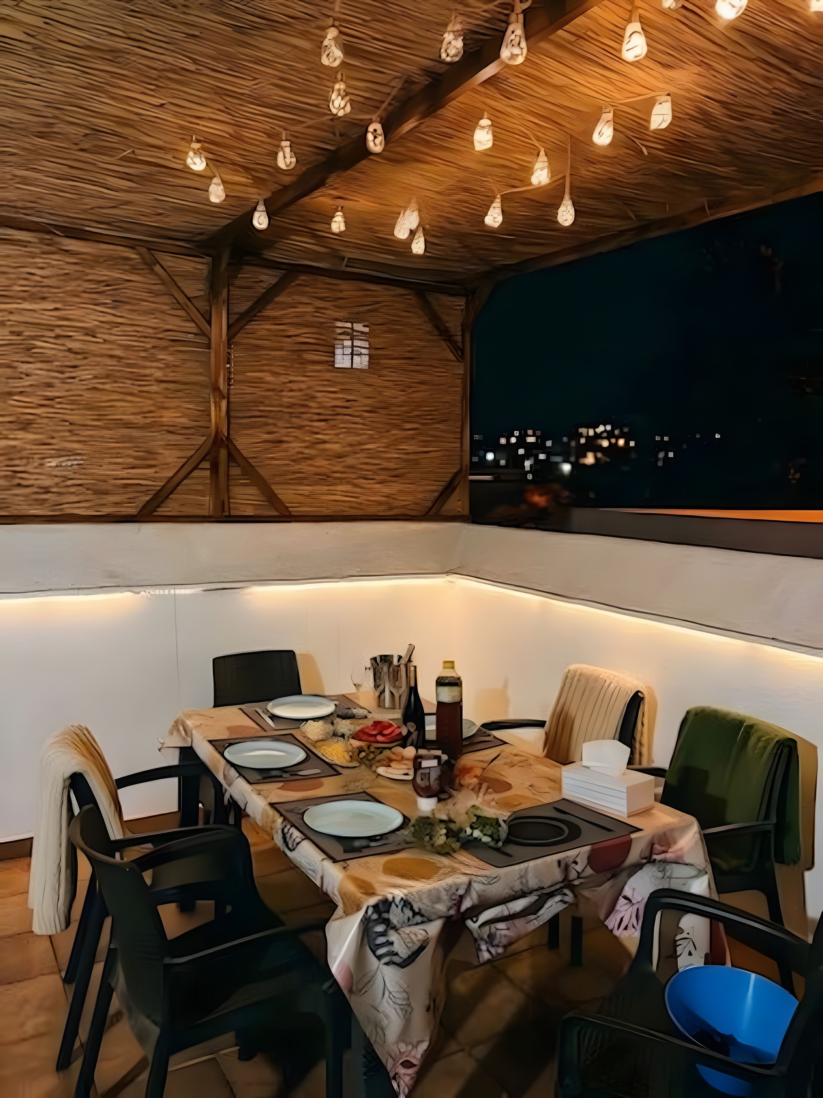 Un área de comedor al aire libre con una mesa dispuesta para cuatro personas, rodeada de sillas, bajo un techo de madera con luces colgantes.