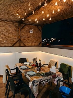 Un área de comedor al aire libre con una mesa dispuesta para cuatro personas, rodeada de sillas, bajo un techo de madera con luces colgantes.