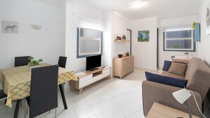 A modern living room with a sofa, a TV, a dining table with chairs, a cabinet, and a small plant. - 2-Bedroom Apartment on Los Cristianos Beach with Promenade Views (Los Cristianos)