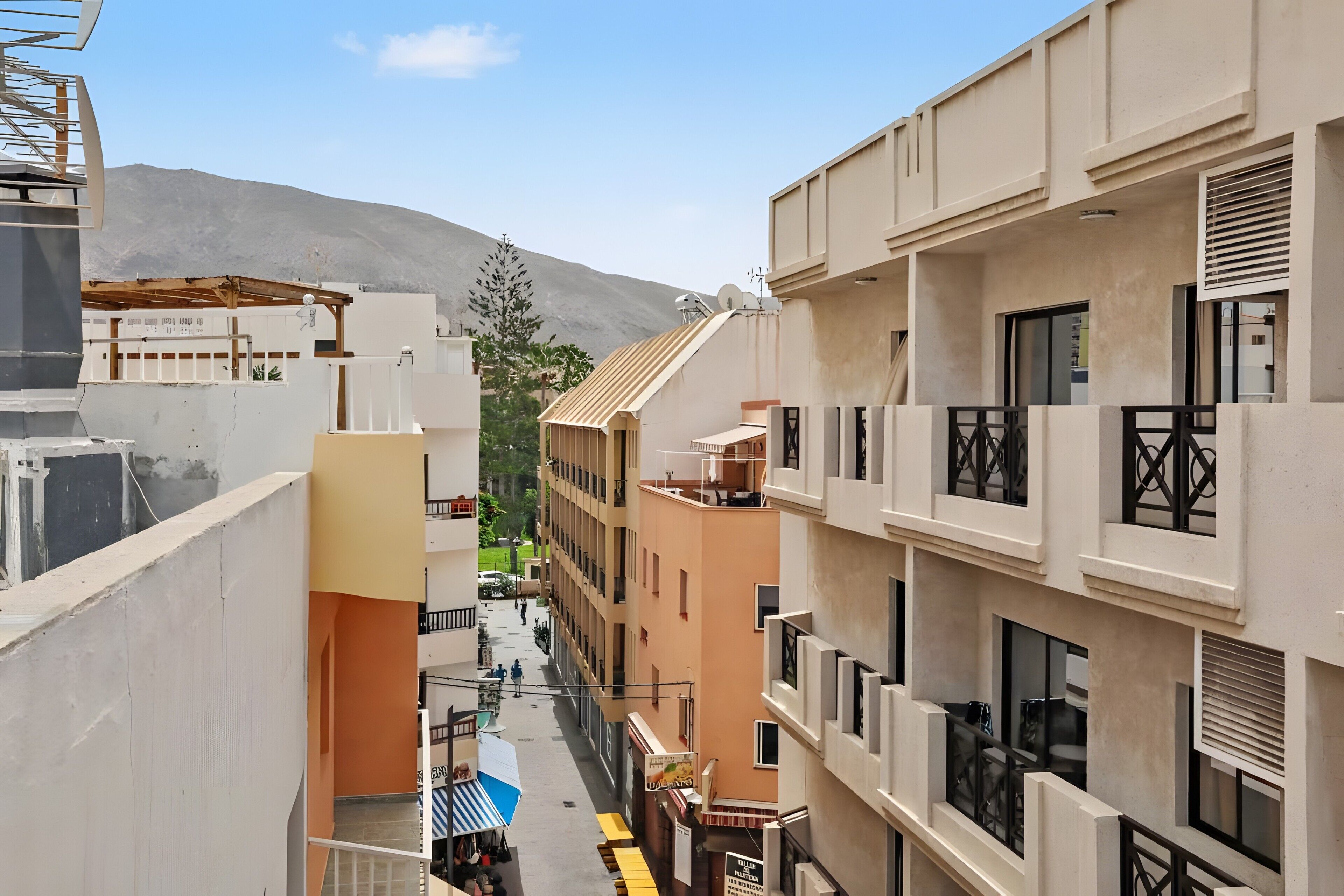 A narrow urban street with multi-story buildings, balconies, and a mountain in the background.