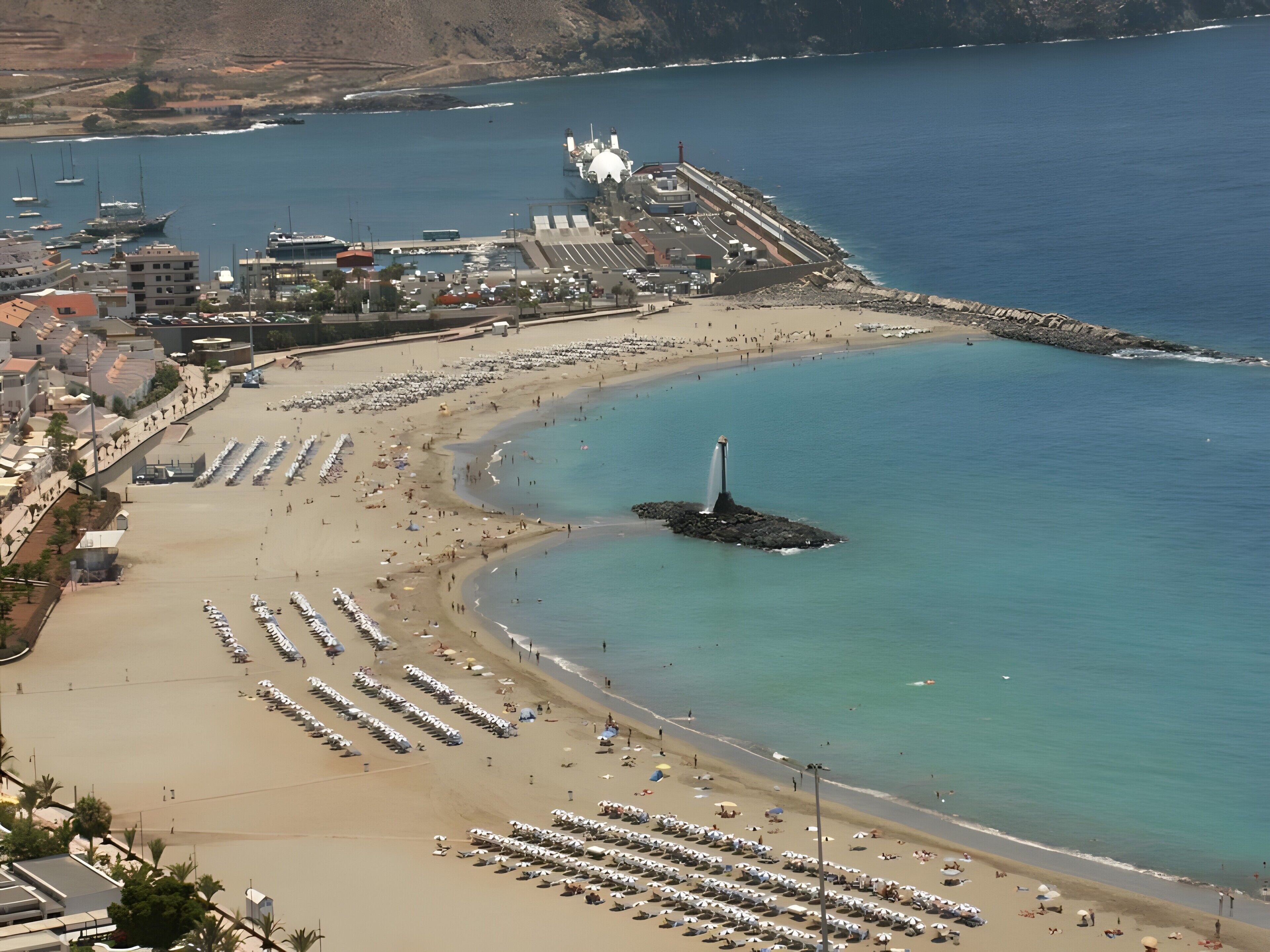 Une plage côtière bordée de rangées de chaises longues et de parasols, un phare et une digue rocheuse.