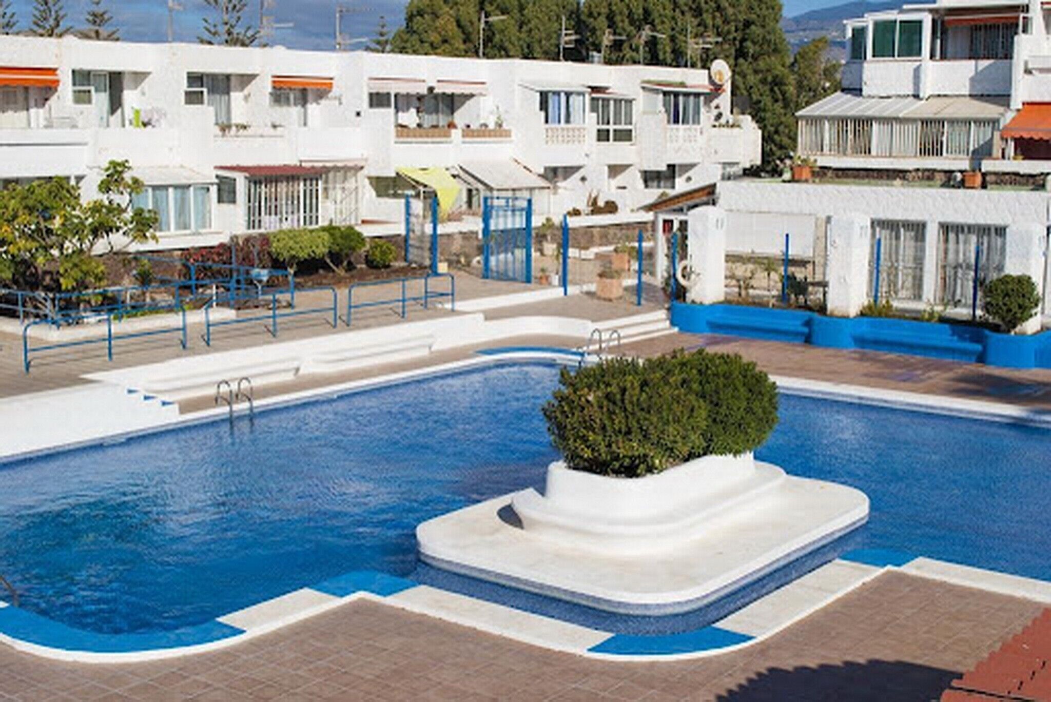 Une piscine avec une petite île au milieu, entourée d’un bâtiment blanc doté de balcons et d’une clôture bleue.