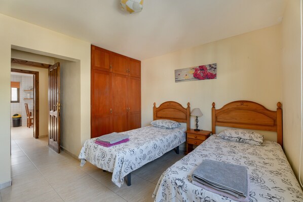 A bedroom with two single beds, wooden wardrobes, a painting on the wall, and a tiled floor.