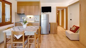 A compact kitchen with wooden cabinets, a stainless steel refrigerator, a dining table with chairs, and a small sofa with a red cushion. - Apartment 'Vittoria Bilo' with Mountain View, Terraces & Wi-Fi (Valfurva)
